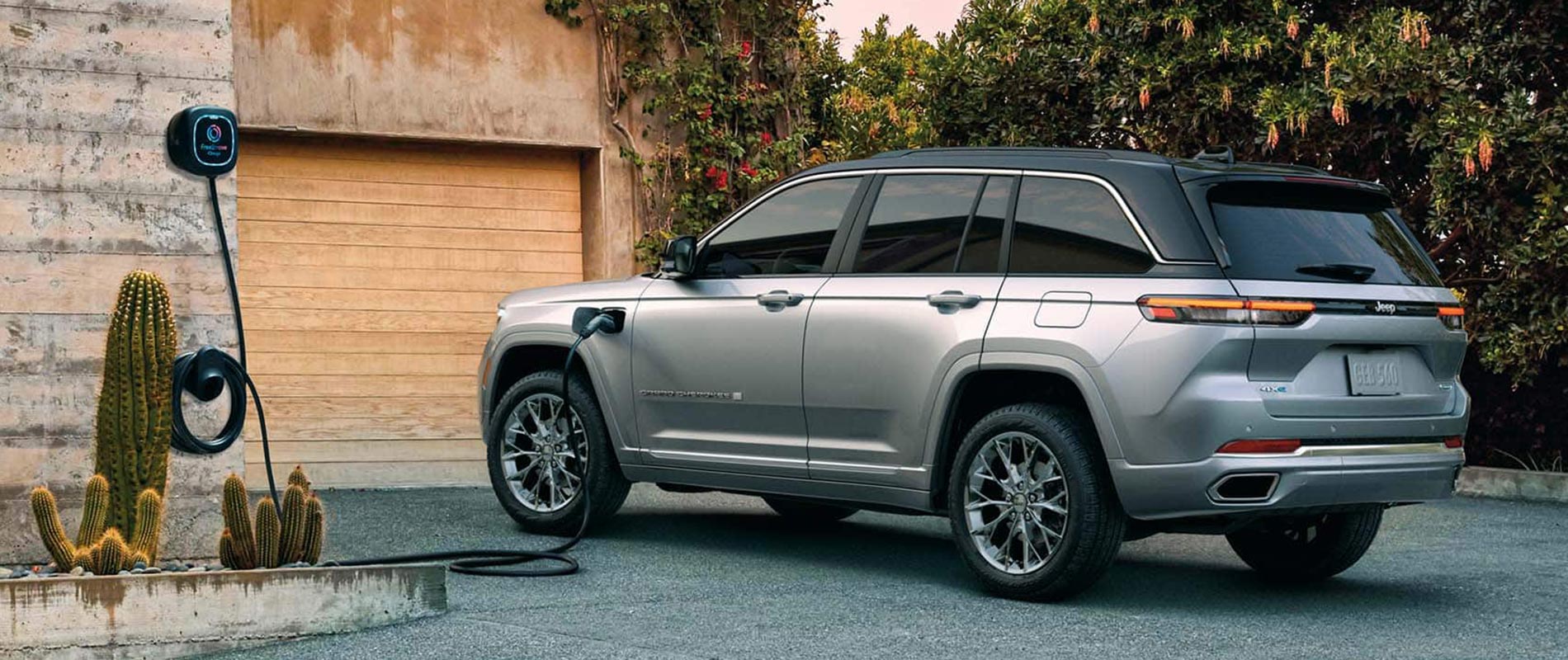 A gray 2025 Jeep Grand Cherokee 4xe parked in a driveway while plugged in and charging.