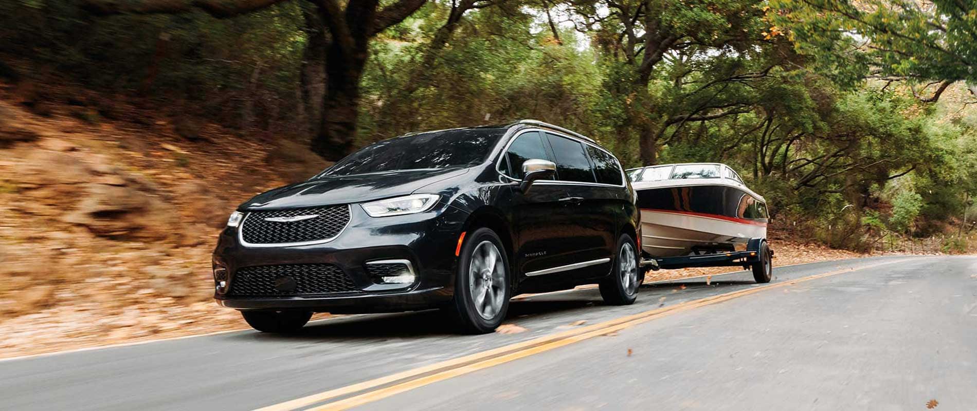 A black Chrysler Pacifica towing a speedboat down a road through a forested area.